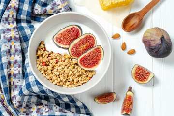 white wheat granola with low-fat yogurt in a white bowl in a composition with a spoon, honeycombs and fig on white wooden background. Healthy breakfast food. Making breakfast. Gluten free