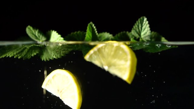 A few cloves of juicy lemon fall into the water through fragrant mint with bunches and splashes on a black background, slow motion, closeup
