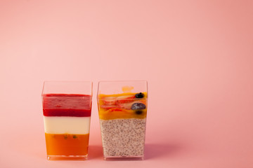 two juicy fresh desserts in cups pudding, chia seeds, mango puree, berries, multi-layer, panakota, passion fruit coconut milk on a pink background in isolated. copy space, place text