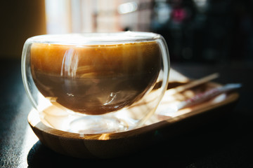 Cappuccino in a transparent glass in the morning sun. Morning in cafeteria. Sunlight. Close up. Coffee mood. Light cup in coffee shop. Sweet and tasty latter macchiato in flare cup on table.