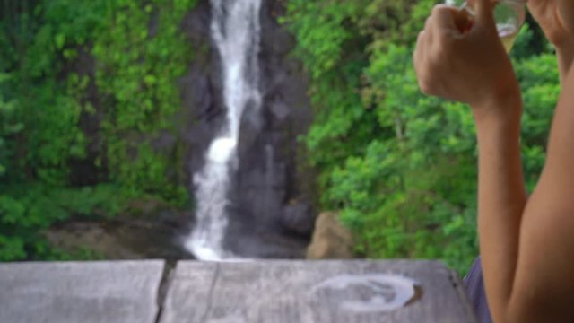 A young woman in a beautiful cafe with an epic view on a canyon and waterfall drinks tea with ice. Travell to Bali concept