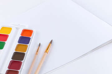 A simple composition of bright color watercolors, two paint brushes and an open album with blank pages for text or picture. Side view, template, mockup.