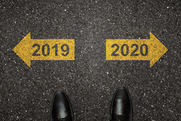 Businessman is looking down at his feet on a Asphalt road with choice arrows and 2019 and 2020 number painted on the surface. Concept for success in the future goal and passing time. Happy new year