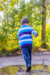 Obraz premium A little boy is jumping in a puddle. A boy in rubber boots. Happy childhood. Puddles after the rain. Warm summer evening.