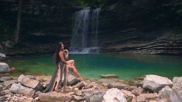 Luxurious attractive brunette woman with long hair in a green dress with bare legs posing in the tropical jungle against the background of a lagoon with waterfall. image of the feminine river nymph