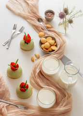 White wooden table with 3 no-bake strawberry mini sphere matcha cheesecakes with bowl of salt caramel, buiscuits, glass pitcher and glass of milk and glass vase with wild teasel flowers.