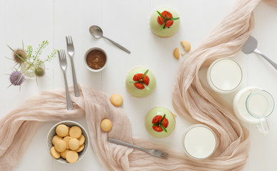 White wooden table with 3 no-bake strawberry mini sphere matcha cheesecakes with bowl of salt caramel, buiscuits, glass pitcher and glass of milk and glass vase with wild teasel flowers.