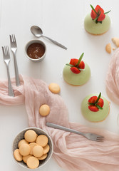 White wooden table with 3 no-bake strawberry mini sphere matcha cheesecakes with bowl of salt caramel and bowl of buiscuits.