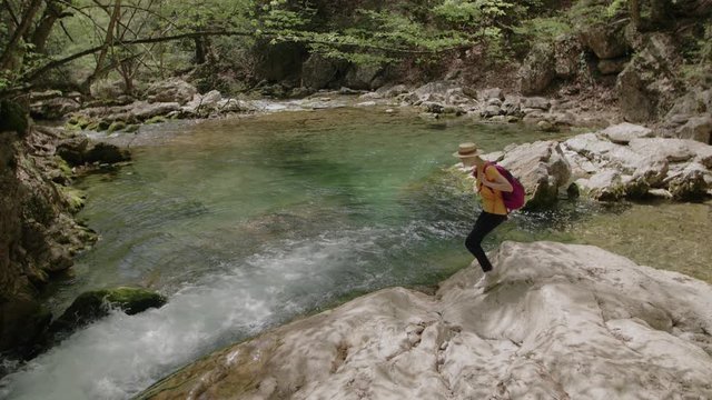 Young tourist woman wearing straw hat, with pink backpack walking to mountain river