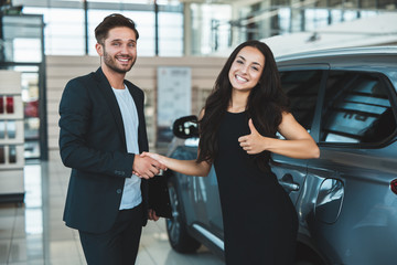 handsome man manager and beautiful brunette woman client shaking hands after succesful deal in dealership center woman showing like standing near brand new car