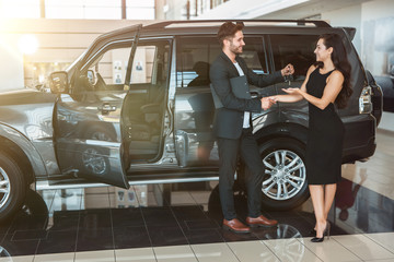 Fototapeta na wymiar handsome man manager gives car keys to beautiful brunette woman client after succesful deal in dealership center standing near new SUV