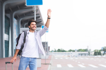 Young guy standing near airport, raising hand and trying to stop car