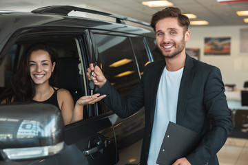 handsome man manager gives car keys to beautiful brunette woman client after succesful retail deal in dealership center standing near new SUV