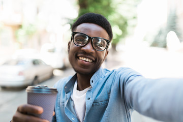 Cheerful young african guy standing outdoors holding coffee make selfie by camera.