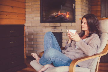 Woman relaxing with cup of tea near fireplace in cozy house.