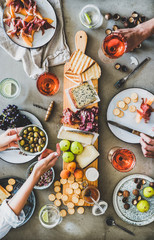 Mid-summer picnic with wine and snacks. Flat-lay of charcuterie and cheese board, rose wine, nuts, olives and peoples hands holding glasses and food over concrete table background, top view