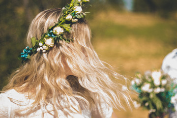 Back of the young woman in a white dress in boho style with a floral wreath in the summer in the field.