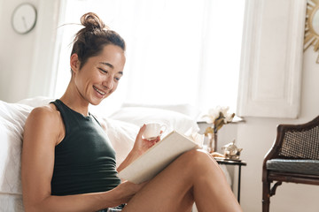 Pleased beautiful woman indoors at home sitting on floor near bed in a bedroom reading book.