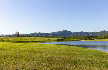 view of green field in front of small lake with moutain and blue sky background