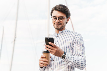 Photo of smiling young man typing on cellphone and drinking coffee from paper cup while walking on pier