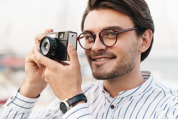 Photo of happy caucasian man taking photo on retro camera and smiling while walking on pier