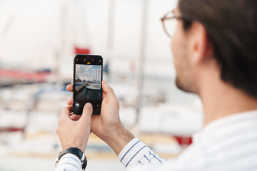 Photo of unshaven brunette man taking photo on smartphone while walking on pier