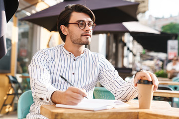 Photo of thinking unshaven man wearing eyeglasses writing on diary and drinking coffee