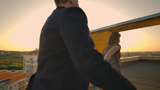 Young couple holding hands walking woman leading boyfriend the roof of the building at sunset POV travel concept. Carefree free lovers run on the roof laughing and smiling.