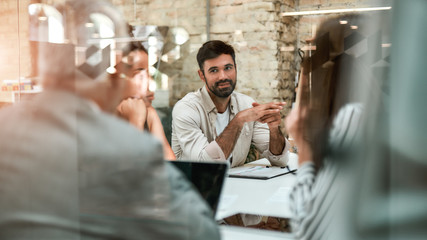 Young business expert. Smiling man discussing something with colleagues while sitting at the office table behind the glass wall
