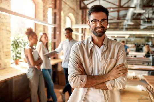 Great working day! Young bearded man in casual clothes keeping arms crossed and looking at camera with smile while standing in the creative office