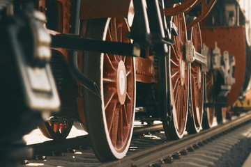A vintage photo of the red big wheels of an old 20th-century locomotive that stands on rails, illuminated by sunlight.