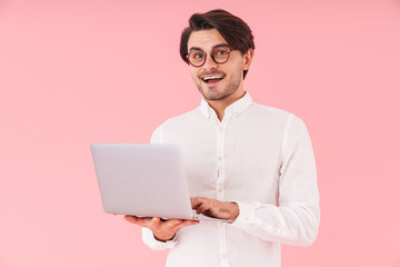 Image of handsome happy man wearing eyeglasses typing on laptop and smiling at camera