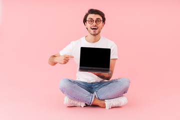 Image of happy handsome man wearing eyeglasses holding and pointing finger at laptop while sitting on floor