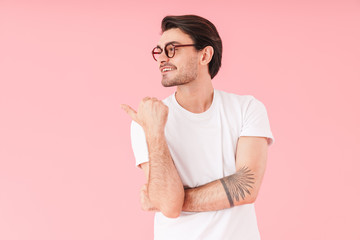 Image of young cheerful man wearing eyeglasses looking and pointing thumb aside at copyspace