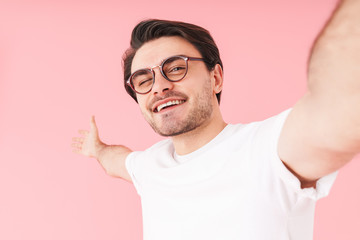 Image of young happy man wearing eyeglasses winking and taking selfie photo