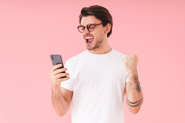 Image of young astonished man wearing eyeglasses holding cellphone and showing winner gesture