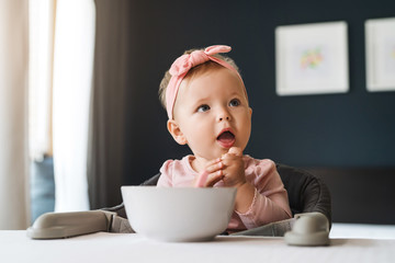 Nine-month-old smiling baby girl sits at white table in highchair and eats herself with spoon from bowl. Blurred background.