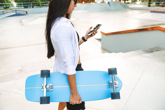 Photo of stylish young woman holding cellphone and carrying skateboard in park