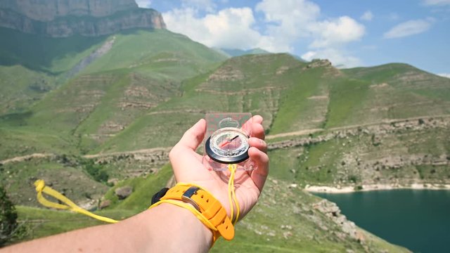 The first-person view of a male hand holding a plastic magnetic compass rotates on the side and looking for the right direction in the background of a mountain. The concept of orienteering