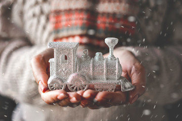 A woman in a beige knitted sweater holds in her hands a Christmas decorative ornament - a silver steam locomotive.
