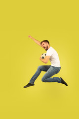 Fototapeta na wymiar Full length portrait of happy jumping man isolated on yellow background. Caucasian male model in casual clothes. Freedom of choices, inspiration, human emotions concept. Playing football on the run.