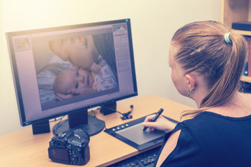 Young woman is editing photos by a graphics tablet and graphics software. Photo of kissing mother of her baby is on computer monitor in front of her. All potential trademarks are removed. 