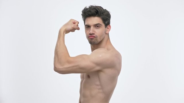 Side view of confident young brunette man with naked torso showing his biceps with raised arm while looking at the camera over white background isolated