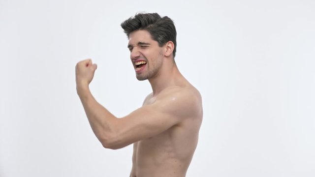 Side view of cheerful young brunette man with naked torso screaming and showing winner gesture with hand while looking at the camera over white background isolated