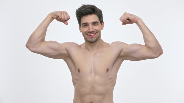 Smiling young brunette man with naked torso showing his biceps and kissing them while looking at the camera over white background isolated