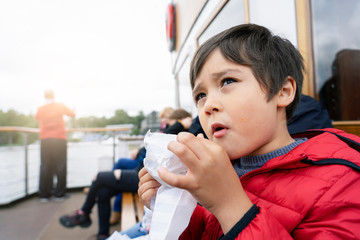 Low view portrait hungry kid eating snack while sitting in boat, cute little boy with dirty face of croissant looking up to sky, Young traveler exploring and learning about the world concept