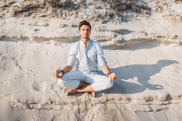 Young handsome man dressed in light clothing sits in meditation pose on sandy beach. Concept of freedom relaxation. Place for text or advertising