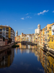 Naklejka na ściany i meble Girona's tipical skyline cityscape over the Onyar River with colourful river houses on a blue sunny sky, Church of Sant Feliu Cathedral landmark on background
