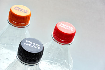 Plastic bottles and orange and red bottle caps with please recycle message against a grey background. Concept of plastic pollution.	