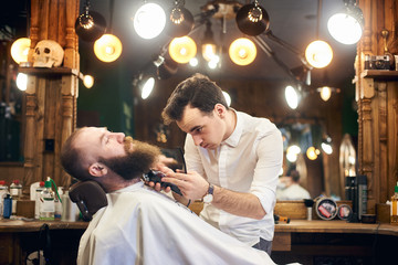 Young concentrated barber shaving and cutting beard with electric trimmer. Fashionable bearded lumberjack style. Barbershop interior with elements of eclecticism and creative lighting. Side view.